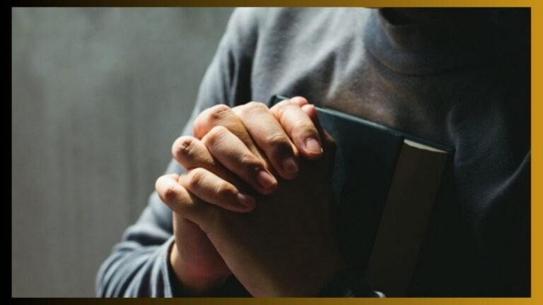 Person’s hands pressed together in prayer while clutching a closed book against a dark background.