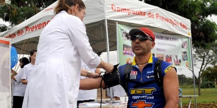 Healthcare worker in a white coat checks a man's blood pressure with a cuff under a 'Campanha de Prevenção' tent at an outdoor health event, park setting.