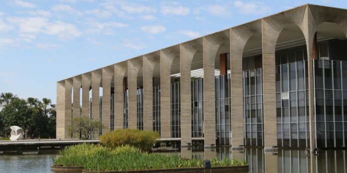 Modern concrete building with tall arched colonnade and glass walls beside a reflecting water feature and greenery.