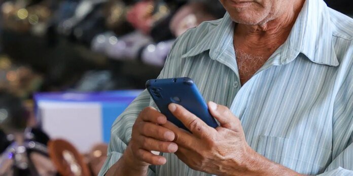 Older man in a light blue striped shirt looking at a blue smartphone, outdoors in a busy setting