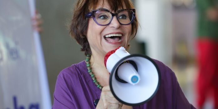 Woman in a purple top and glasses smiles while speaking into a red and white megaphone.