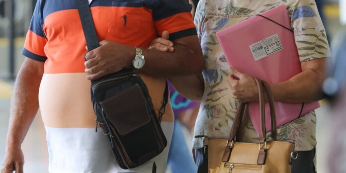 Two adults walk together, arms linked; man wears a striped orange polo with a crossbody bag and watch, woman holds a pink folder and tan handbag.