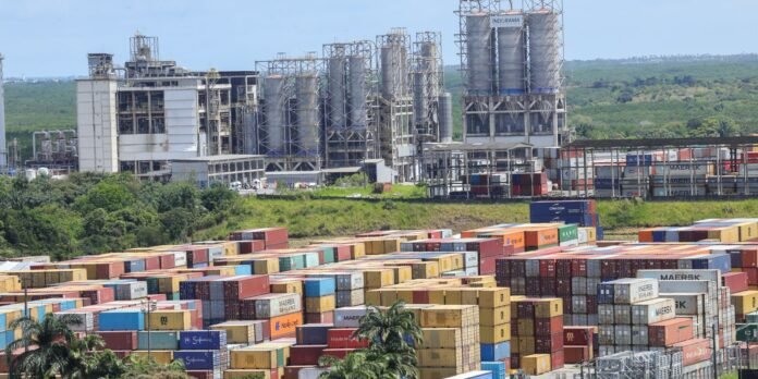 Colorful shipping containers fill a port yard with a large industrial facility and silos rising in the background.