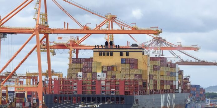 Container ship at a busy port loaded with stacked colorful shipping containers, with orange gantry cranes towering above the harbor.