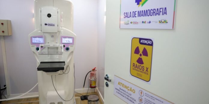 Mammography machine in a clinical room with control panels and a chest plate, next to a fire extinguisher and warning signs on the wall.