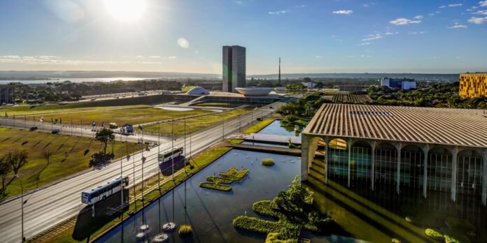 Aerial view of a modern city center with a tall twin-tower building, a white domed pavilion, and a reflecting pond with island greenery.