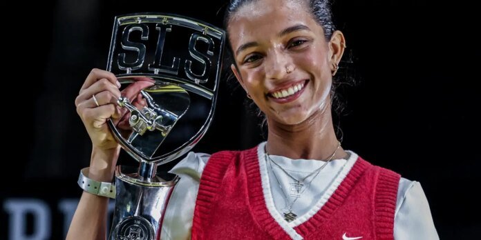 Smiling female athlete holding a large silver trophy with 'SLS' letters, wearing a red sleeveless sweater over a white shirt.