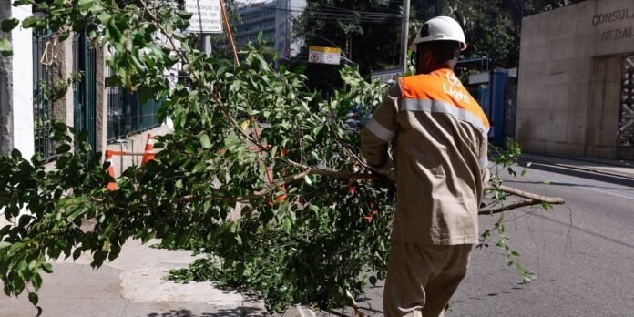 Worker in orange safety vest and hard hat clears a large fallen branch from a city street sidewalk area.
