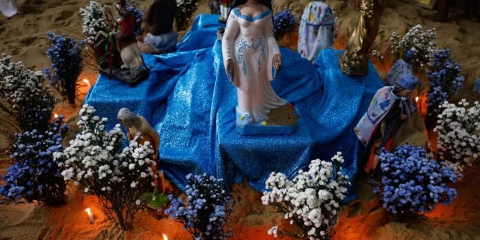 Nativity scene featuring statues on a blue glittery cloth with white and blue flowers and small candles around the sand-hued ground.