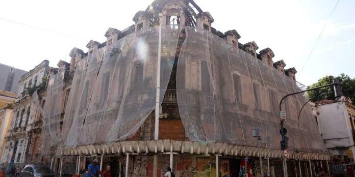 Historic multi-story building wrapped in construction netting on a city street, with scaffolding and pedestrians nearby