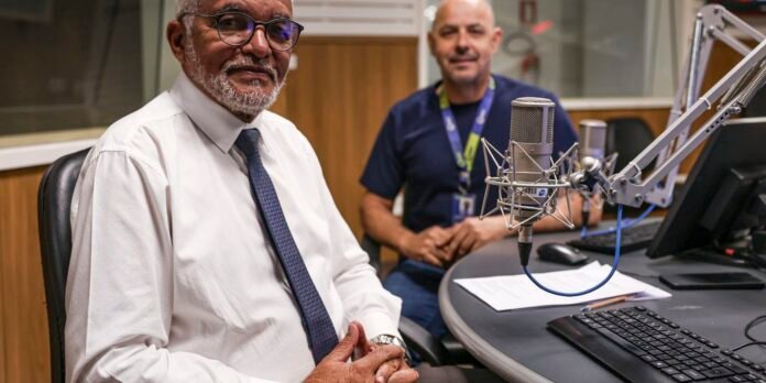 Two men sit in a radio studio with a large studio microphone on a boom arm between them; one in a white shirt and tie speaks while the other looks on.