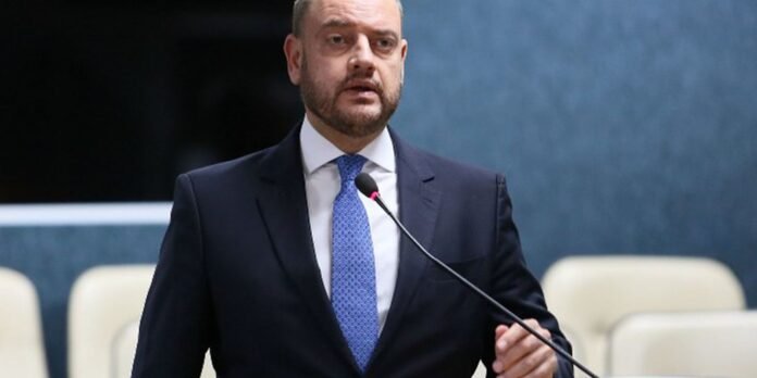 Man in a dark suit and blue tie speaks into a microphone at a formal event.