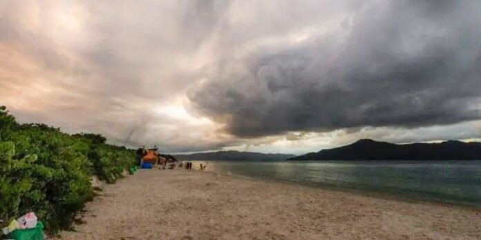 Coastal beach scene with dark storm clouds over the water, a lifeguard chair near the shore, and people on the sandy beach; greenery on the left.