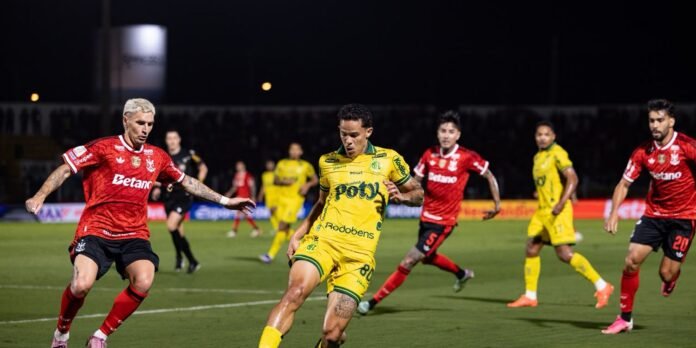 Soccer player in yellow lunges to kick the ball while red-clad opponents close in during a nighttime match on a green field
