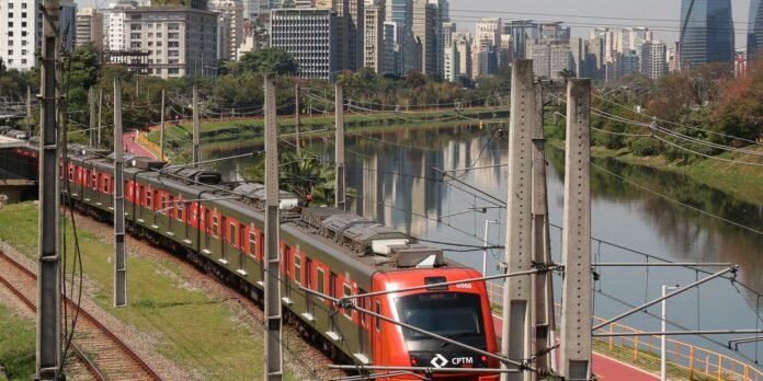 Red and green passenger train weaving along a riverbank with a city skyline in the background.