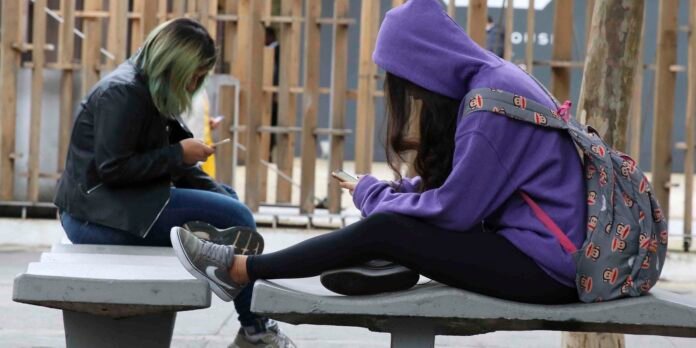 Two young people sitting on concrete benches outdoors, both looking at their phones; one wears a purple hoodie and a monkey-print backpack.