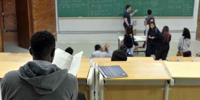 Student in a gray hoodie reads a notebook at a university desk, with a chalkboard and groups of students in the background.