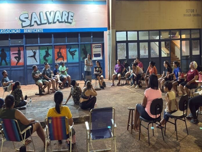 Community meeting in a covered outdoor space with a speaker standing while attendees sit in a circle. The wall behind shows colorful silhouettes and a 'SALVARE' sign.