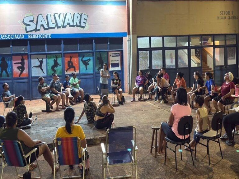 Community meeting in a covered outdoor space with a speaker standing while attendees sit in a circle. The wall behind shows colorful silhouettes and a 'SALVARE' sign.