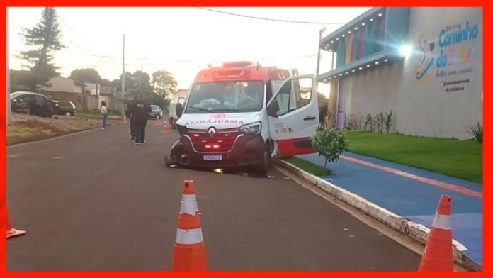 Ambulance with front-end damage stopped on a street, orange cones around it, door open near a colorful storefront.
