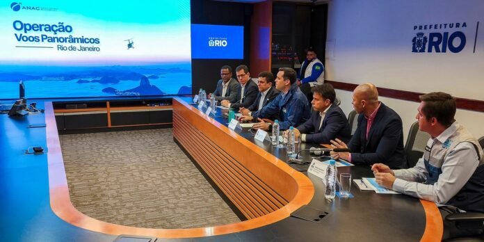 Panel of officials seated at a curved conference table during a briefing, with a large screen displaying 'Operação Voos Panorâmicos Rio de Janeiro' in the background