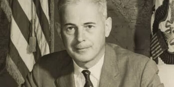 Portrait of a man in a suit and tie with flags in the background.