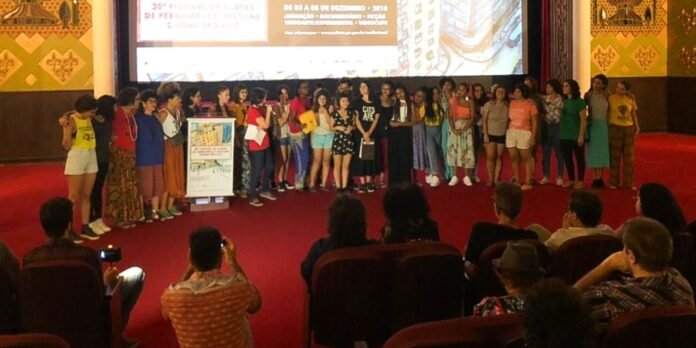 Group of people on a red-carpet stage posing for a presentation, with a podium and a large screen behind them.