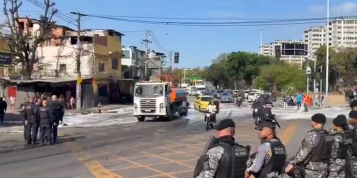 Armed police in tactical vests stand on a busy city street as a white truck passes by and bystanders watch.