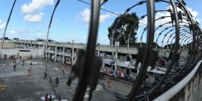 Open-air prison yard with concrete dormitories, laundry hanging outside the cells, and people playing on the dusty ground; barbed wire coils in the foreground.