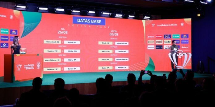 Stage at a Copa do Brasil event with a large red screen displaying match groups and dates, presenter at left podium and audience in foreground