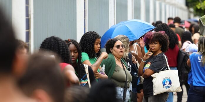 Diverse group of people standing in a long outdoor queue along a fence, some with bags and an umbrella visible in the middle.