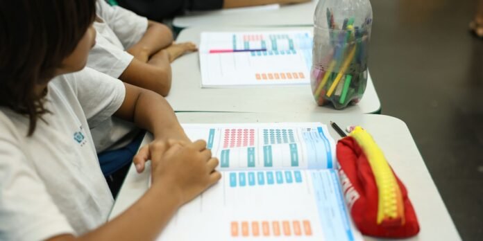 Students at a desk reading a workbook with colorful charts; pencil case and a bottle with pens nearby.