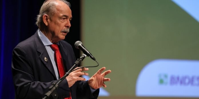 Older man in a dark suit and red tie speaks into a microphone at a conference podium.