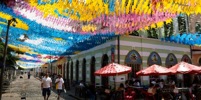 Street scene with vibrant pennant banners overhead, outdoor cafe seating under red umbrellas, and people walking along the cobblestone street.