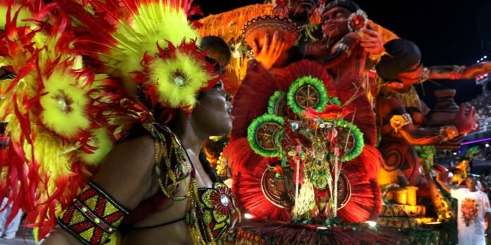 Carnival performers in bright feather headdresses and elaborate beadwork on a decorated parade float at night.