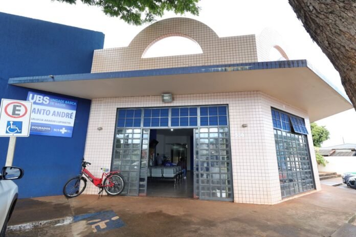 Exterior view of a small health clinic with blue wall, beige tiled facade, and glass block doors; signs indicate a basic health unit and wheelchair accessibility; a red bicycle is parked outside.