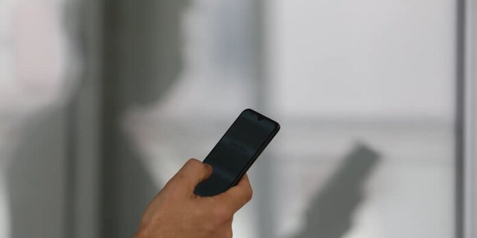 Close-up of a hand holding a dark smartphone indoors, the screen off and a blurred background visible.