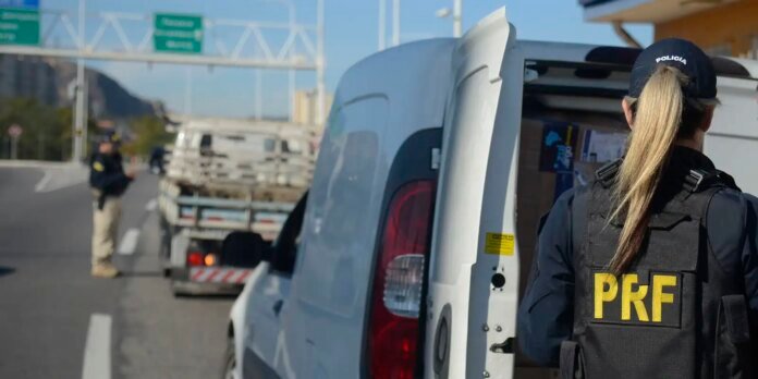 Brazilian PRF officer in a black vest with 'PRF' on the back stands at a highway checkpoint by a white van, with a tow truck in the background.