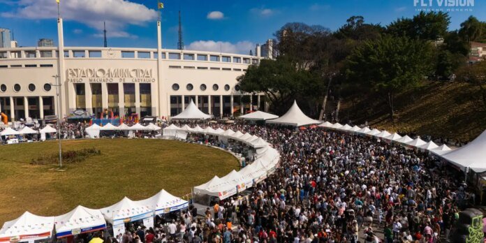 Crowd of festival-goers outside Estádio Municipal Paulo Machado de Carvalho, with white tents lining a curved walkway in front of the stadium.