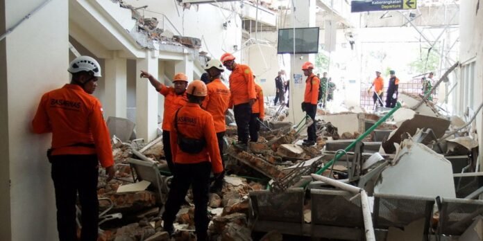 Rescue workers in orange uniforms and helmets survey a collapsed building with rubble and debris all around.